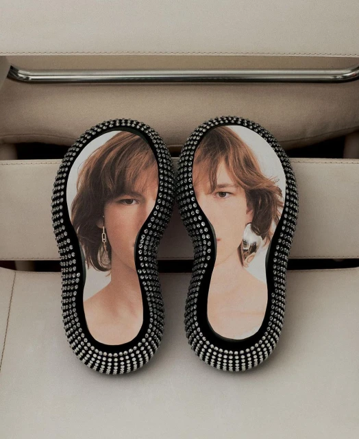 Unique pair of black platform sandals adorned with rhinestones, featuring a model's portrait on each shoe, resting on an off-white surface.