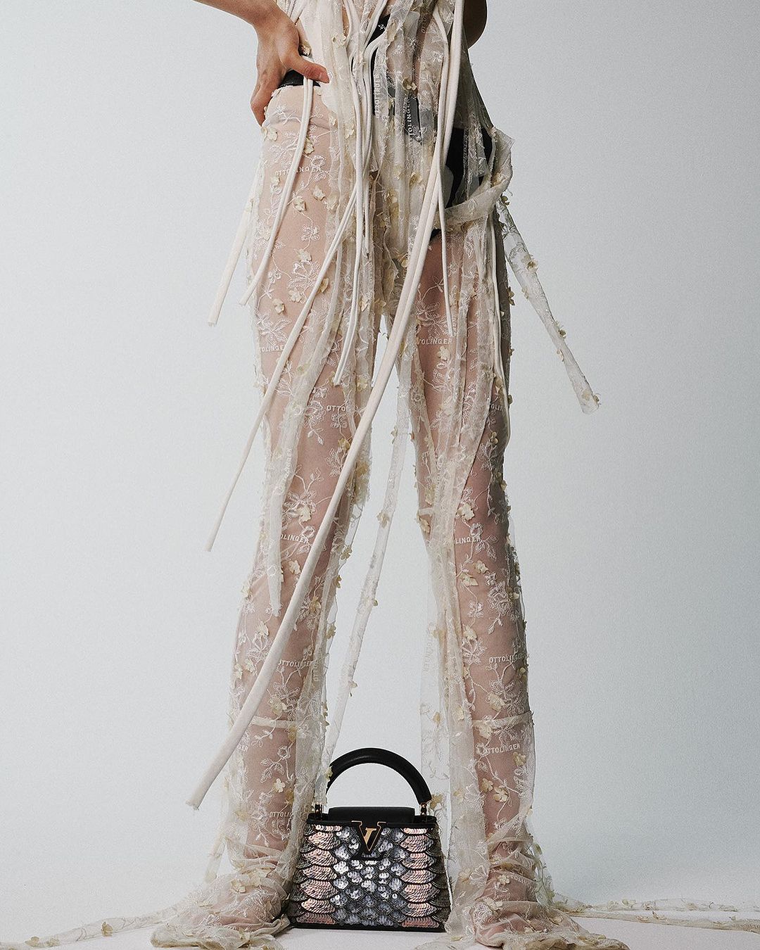 Model wearing a lace ensemble with a black and pink handbag, posing in a bright, white studio setting with a shallow depth of field.