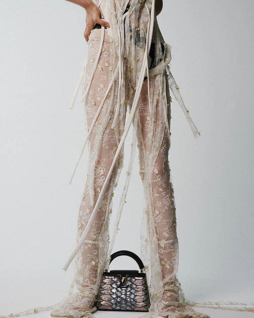 Model wearing a lace ensemble with a black and pink handbag, posing in a bright, white studio setting with a shallow depth of field.