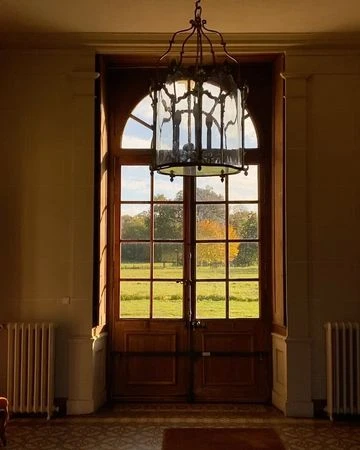 Interior view through a window, showcasing the light fixture, architectural details, and a vibrant landscape of trees and sky beyond.