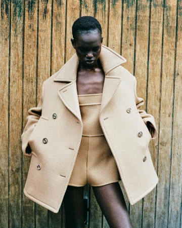 Model wearing an oversized camel wool peacoat and matching shorts against a rustic wooden paneled background in Paris