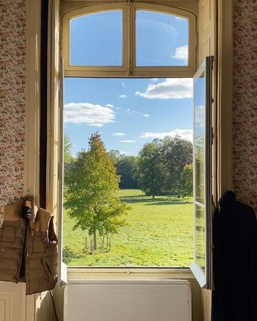 A serene view through an open window of a lush green field dotted with trees under a bright blue sky with fluffy white clouds.