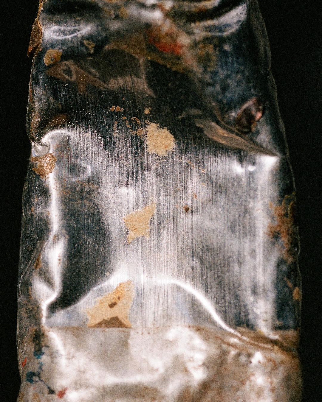 Close-up studio shot of a weathered, metallic tube, featuring scratches and traces of ochre and red paint against a dark backdrop.