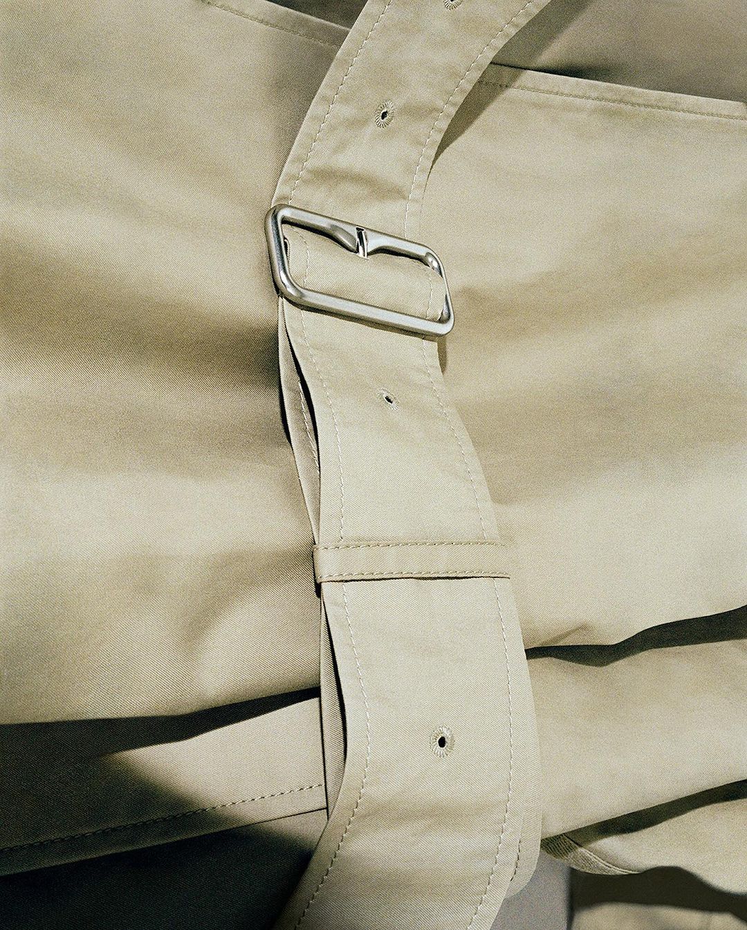 Close-up of a beige trench coat with a prominent buckle. Focus on the garment's details, texture, and the play of light and shadow.
