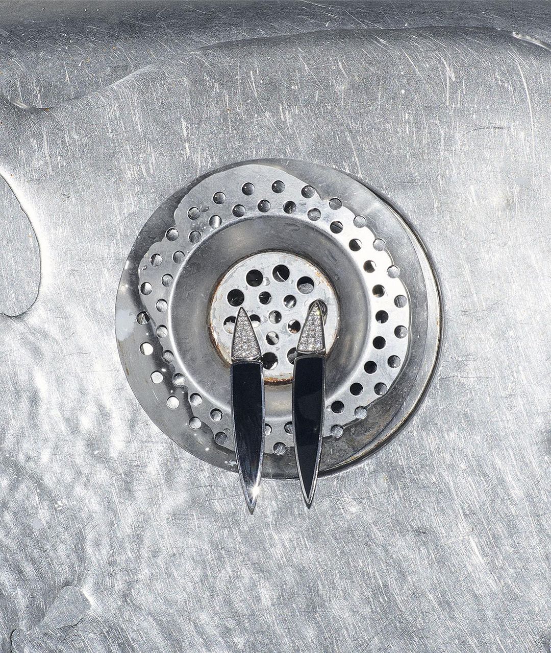 Close-up of two black and silver earrings with diamond accents placed on a stainless-steel sink drain in a photo studio setting.