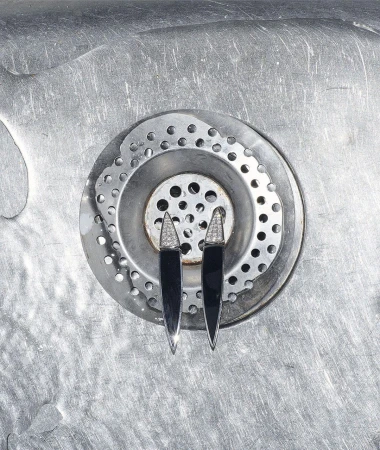 Close-up of two black and silver earrings with diamond accents placed on a stainless-steel sink drain in a photo studio setting.