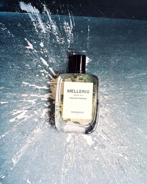 Mellerio perfume bottle on a speckled surface with white radial paint splatters, close-up studio shot, product photography.