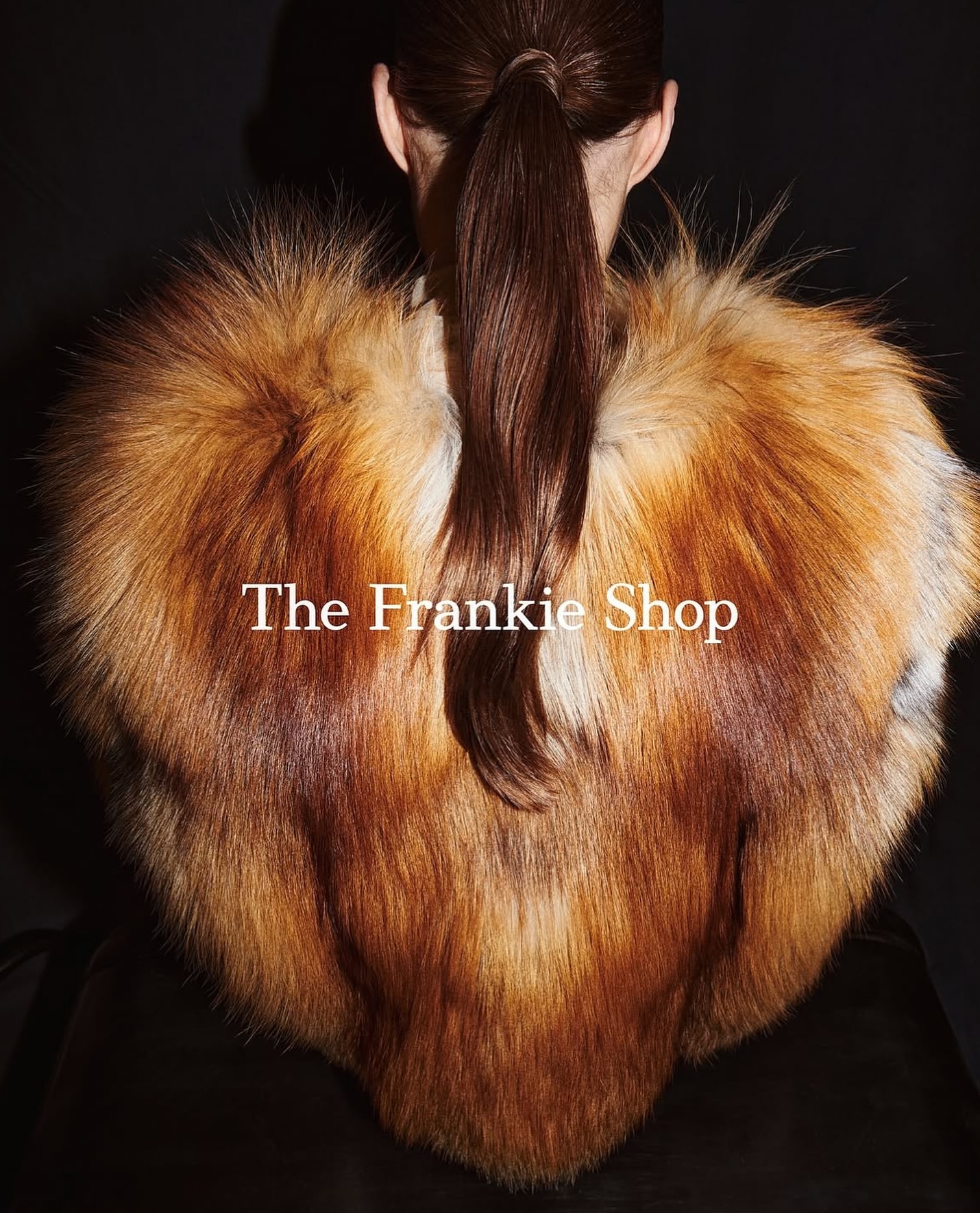 Back view of a woman with brown hair in a ponytail wearing a large brown and orange fur shawl against a dark background.