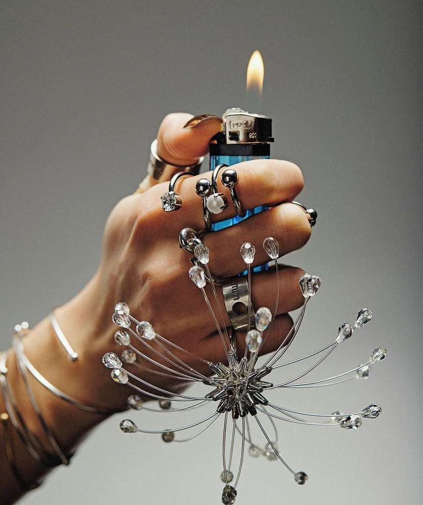 Hand adorned with rings and bracelets holding a lit lighter with a decorative wire embellishment, set against a plain grey background.