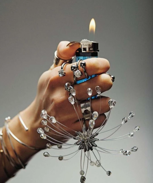 Hand adorned with rings and bracelets holding a lit lighter with a decorative wire embellishment, set against a plain grey background.