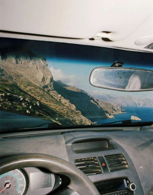 Car interior view of a rocky coastal landscape visible through the windshield, featuring mountains and blue sea.