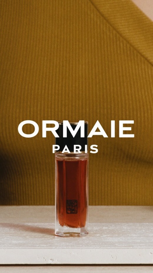 Elegant perfume bottle stands on a surface with a blurred figure wearing an olive-green top in the background, product shot.
