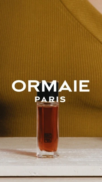 Elegant perfume bottle stands on a surface with a blurred figure wearing an olive-green top in the background, product shot. 