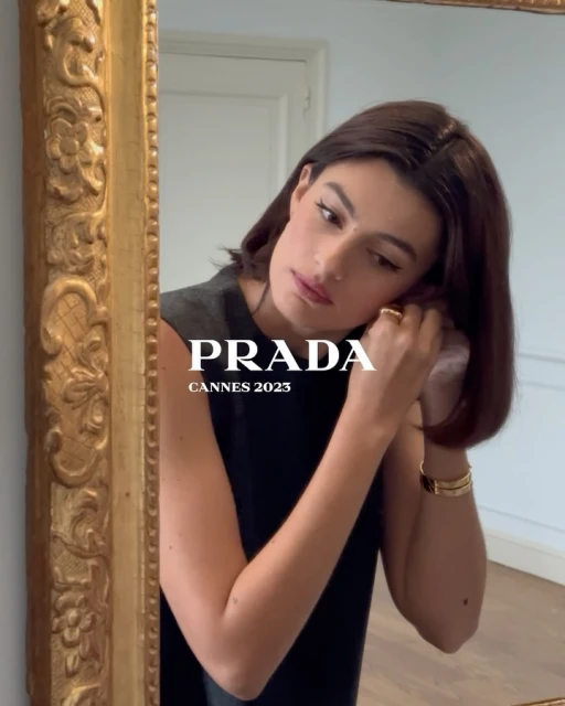 Brunette woman adjusts an earring while looking in a gold-framed mirror inside an indoor setting with natural lighting.