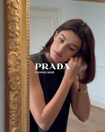 Brunette woman adjusts an earring while looking in a gold-framed mirror inside an indoor setting with natural lighting.