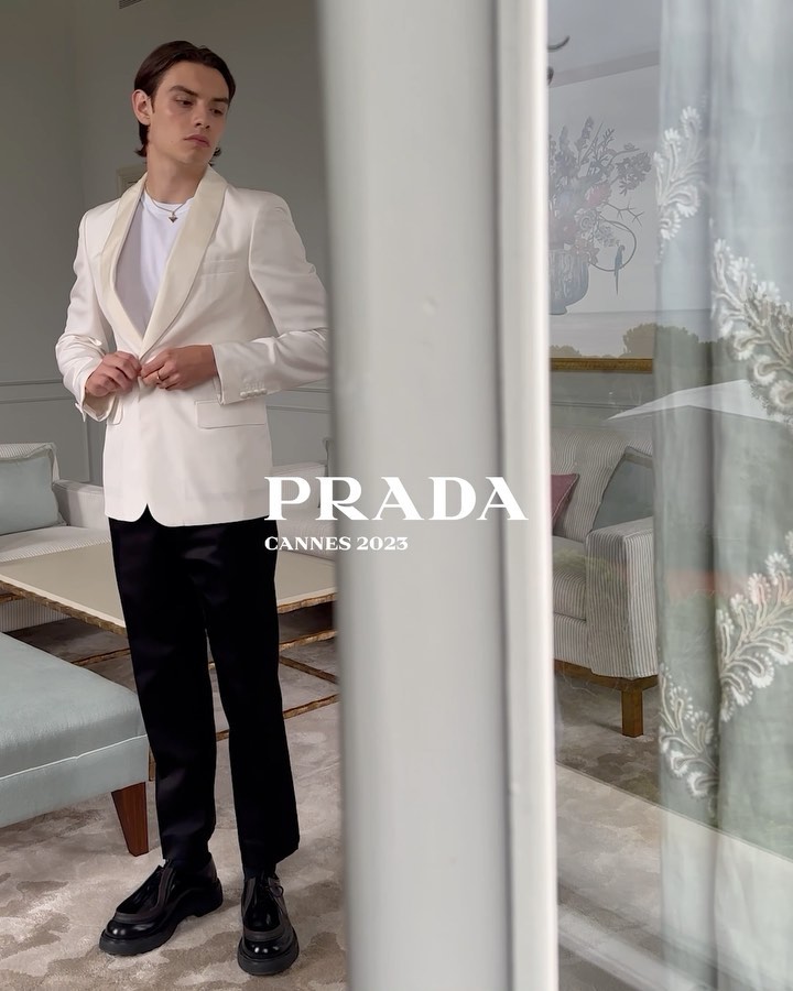 Young man in a white tuxedo-style jacket and black pants strikes a full-length pose indoors near a bright window with PRADA logo text overlay.
