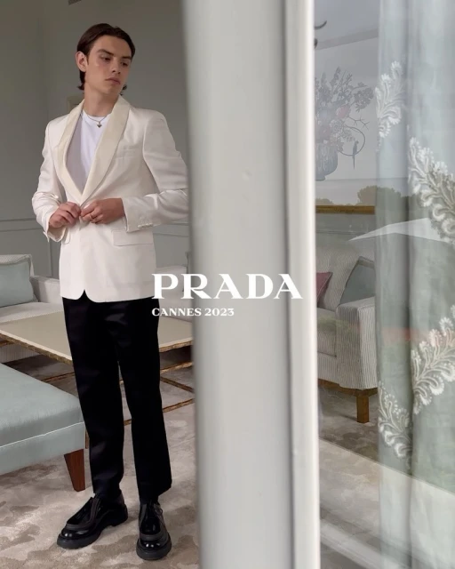 Young man in a white tuxedo-style jacket and black pants strikes a full-length pose indoors near a bright window with PRADA logo text overlay.