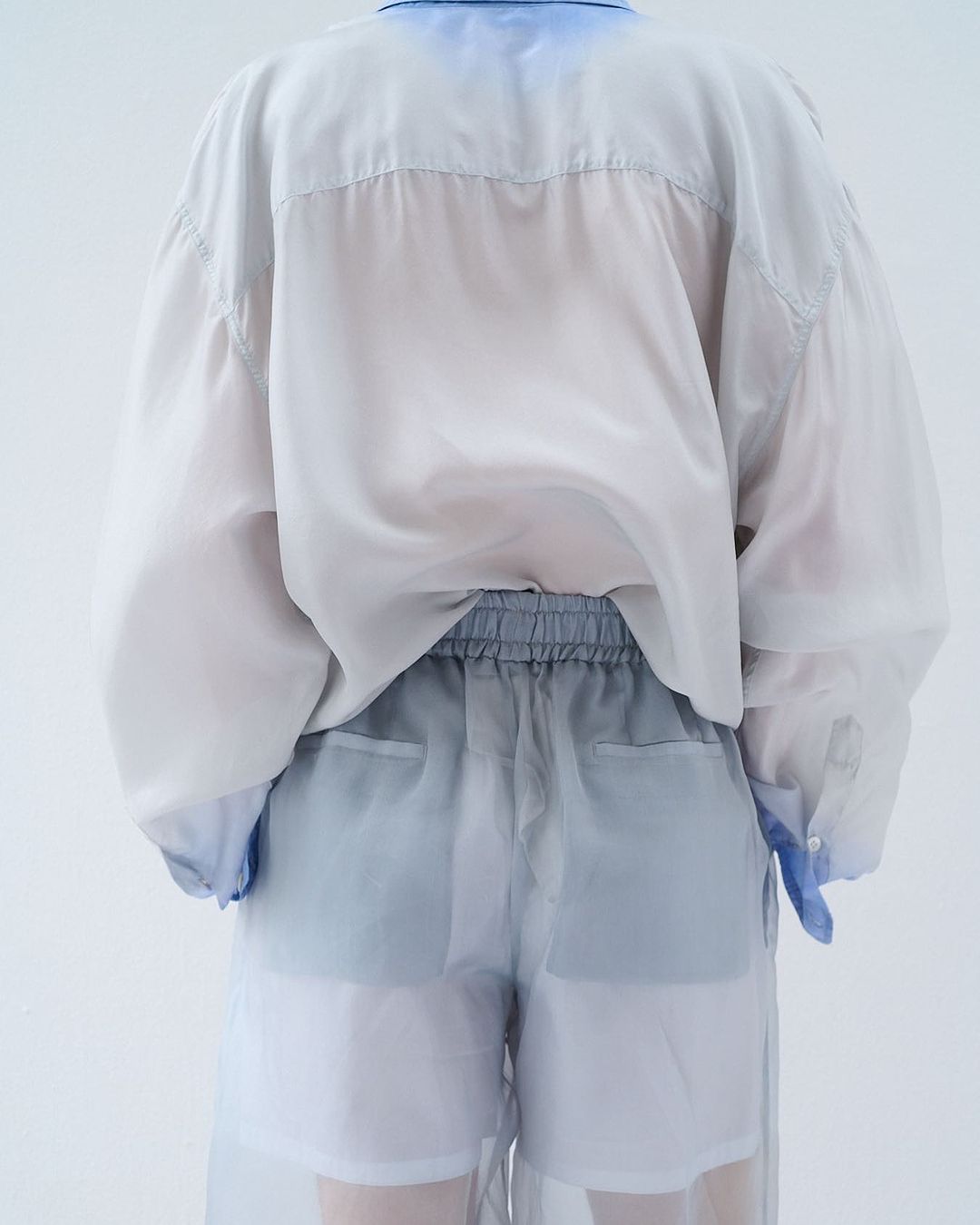 Back view of person wearing a sheer white button-down shirt and sheer shorts with blue-grey accents in a white studio setting.