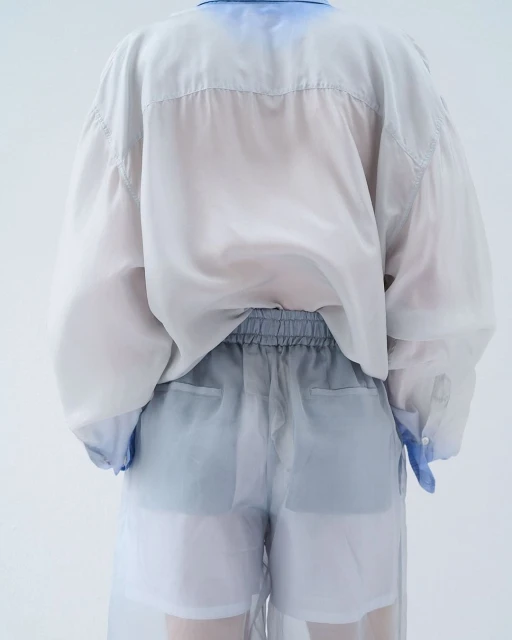 Back view of person wearing a sheer white button-down shirt and sheer shorts with blue-grey accents in a white studio setting.