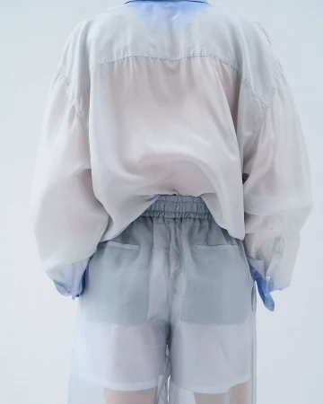 Back view of person wearing a sheer white button-down shirt and sheer shorts with blue-grey accents in a white studio setting.