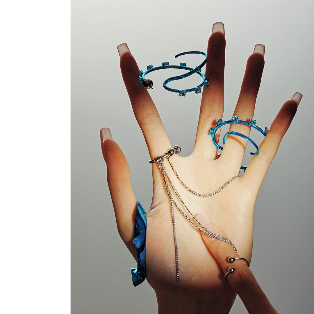 Hand adorned with blue and silver rings and chain details against a light grey backdrop in a studio fashion shot.