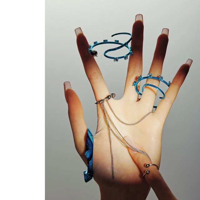 Hand adorned with blue and silver rings and chain details against a light grey backdrop in a studio fashion shot.