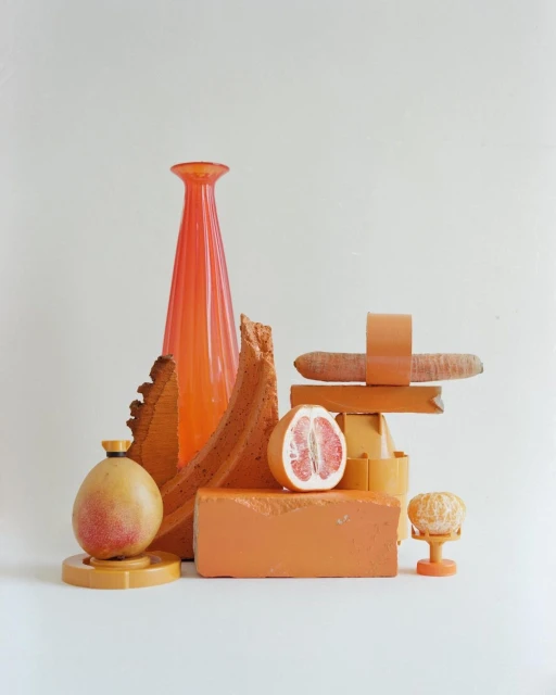 Still life composition featuring a vase, grapefruit half, mango, carrot, and construction materials arranged against a white background.