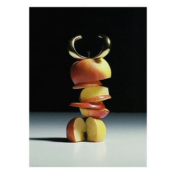 An artistic still life photograph features a stack of sliced apples topped with a gold bracelet against a dark background.