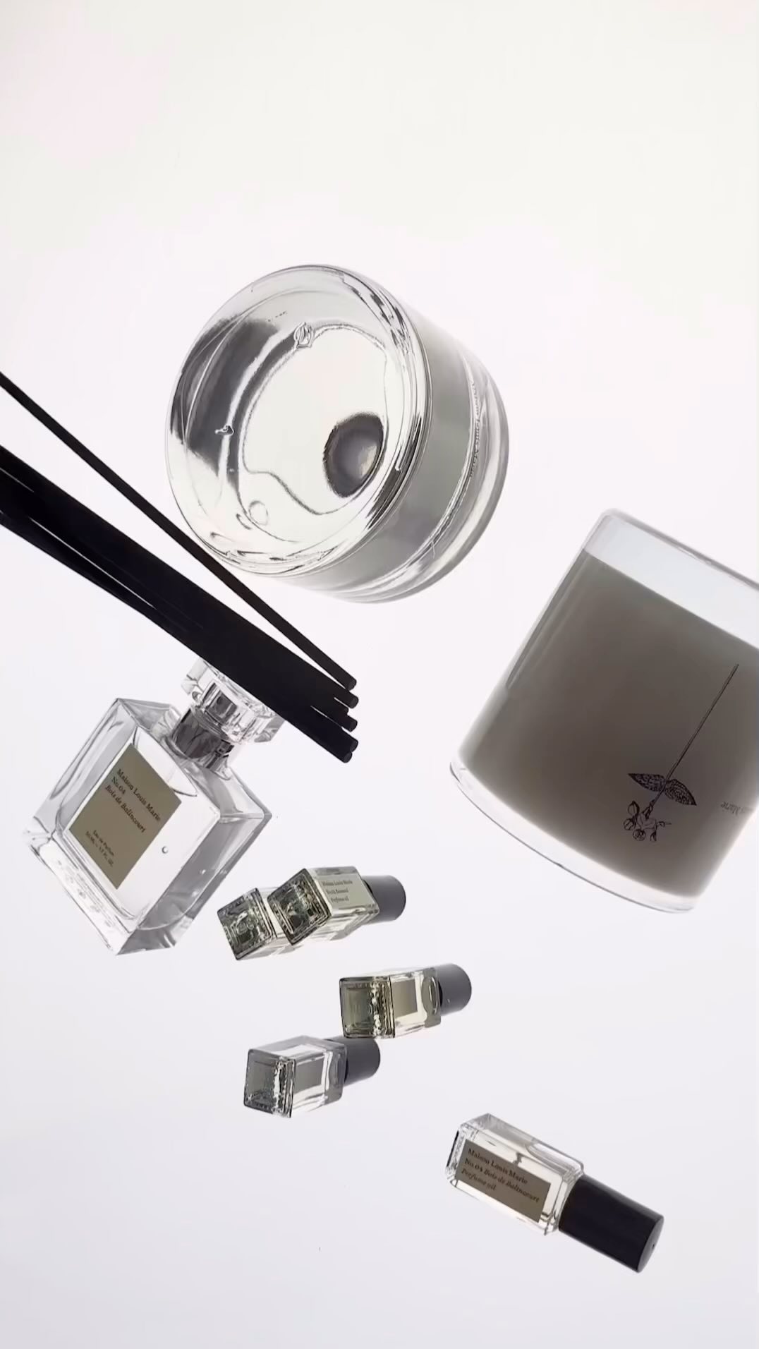 Aesthetic floating shot of a candle, small perfume vials, and scent diffuser with dark reeds against a plain white backdrop in a still life.