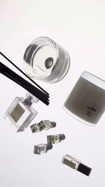 Aesthetic floating shot of a candle, small perfume vials, and scent diffuser with dark reeds against a plain white backdrop in a still life.