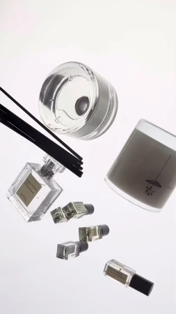 Aesthetic floating shot of a candle, small perfume vials, and scent diffuser with dark reeds against a plain white backdrop in a still life.