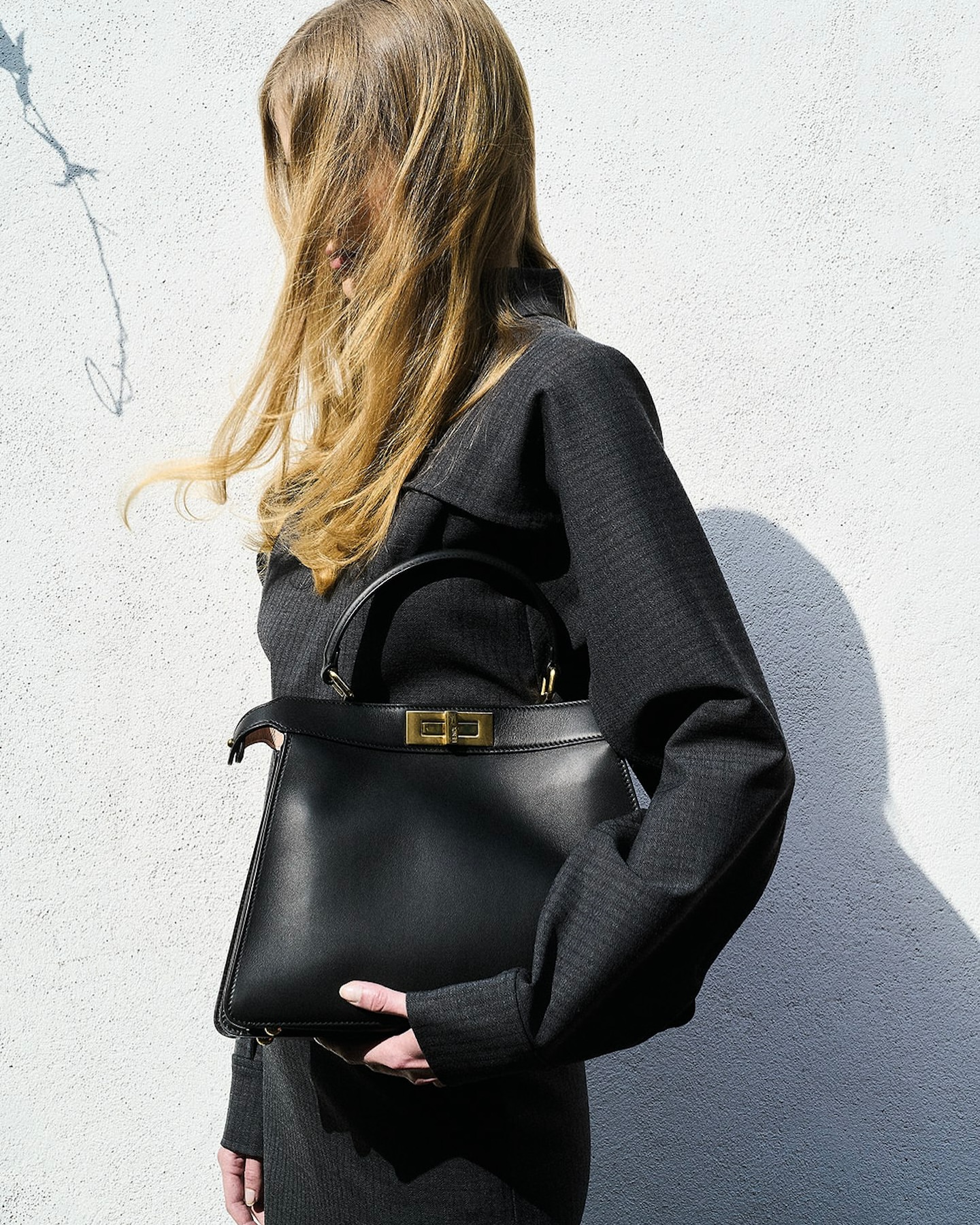 Woman in gray outfit holding black leather handbag in studio. Fashion shot of purse with hand model in front of white wall.