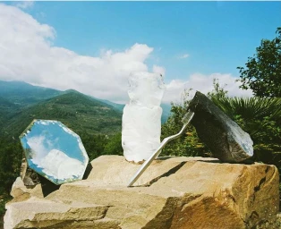 A toothbrush, a faceted mirror, an ice sculpture, and a dark stone resting on a sunlit rock against distant green mountains.