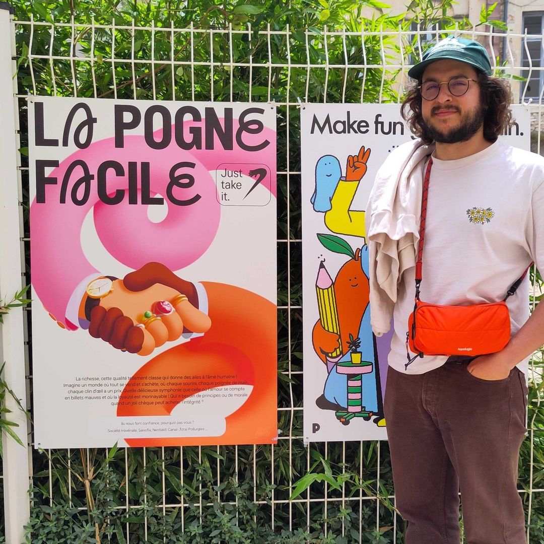 Man stands in front of posters on a white fence with green foliage. He wears a hat, t-shirt, and orange crossbody bag.
