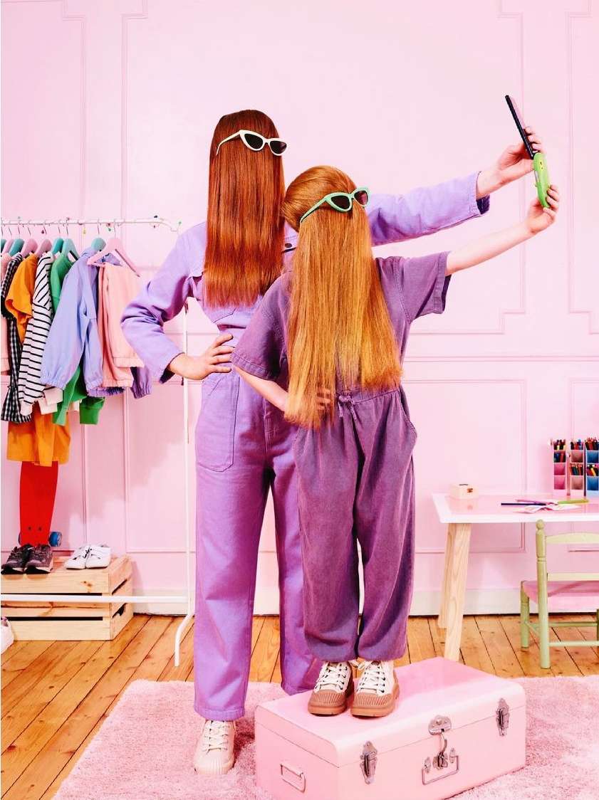 Two red-haired subjects in lilac outfits pose in a pink interior. A girl stands on a pink trunk, holding a phone up for a selfie.