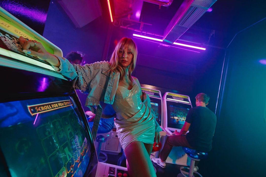 Young woman poses leaning on vintage arcade game, in short dress and beaded denim jacket, at an entertainment center with neon lights.