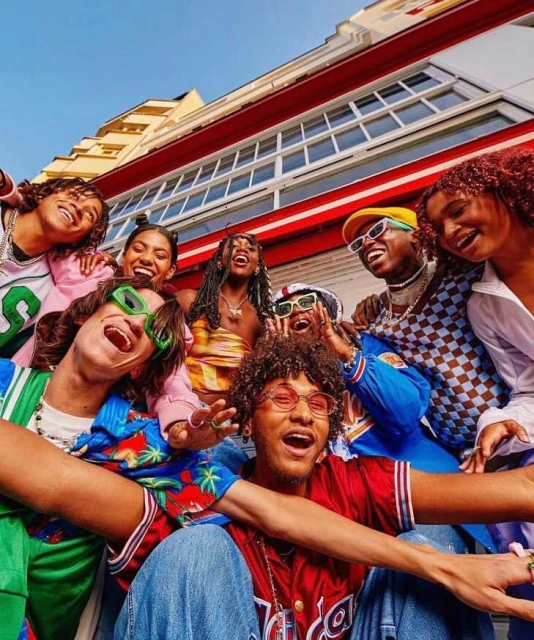 Group of diverse young adults smiling and posing together outdoors in casual streetwear with an urban background on a sunny day.