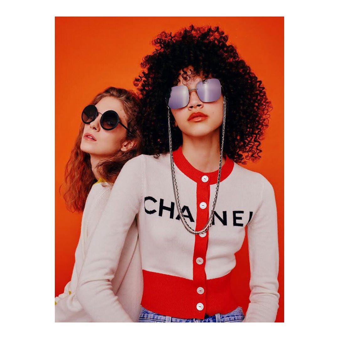 Two young adult women pose in front of an orange background. One woman wears round dark glasses, the other wears silver square glasses.