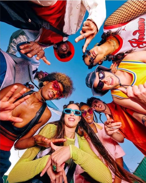 A diverse group of young adults in colorful clothing gather, looking down at the camera with sunglasses on, in an eye-level angle shot against a blue sky.