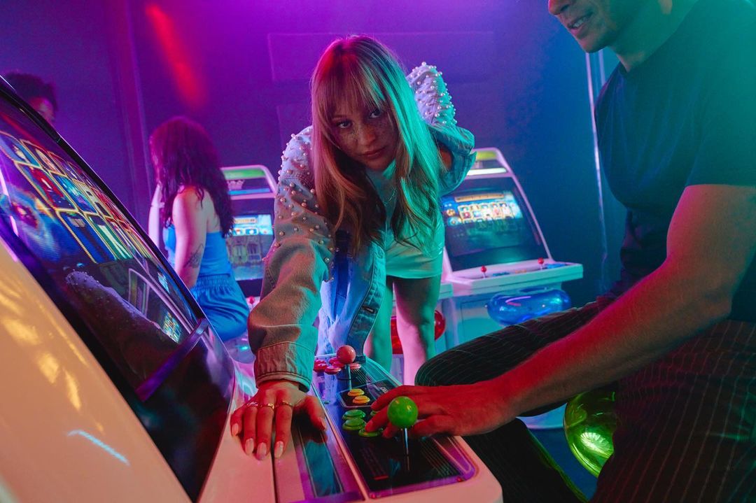 People play a classic arcade game in a neon-lit game room. A woman is in focus and presses buttons on the arcade game, other people in background.