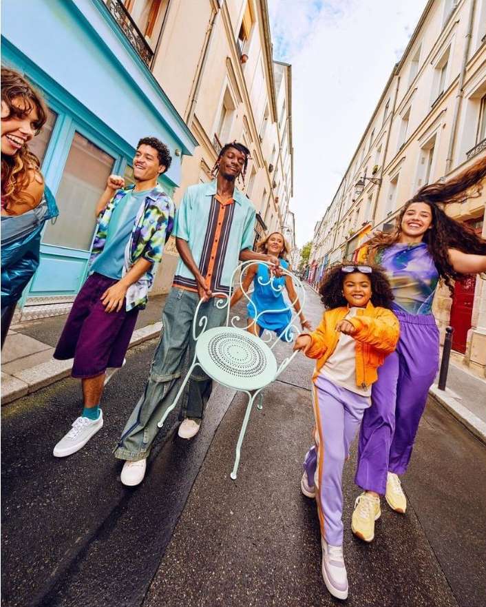 A group of diverse young adults walking down a narrow city street, wearing colorful casual clothing, holding a small chair, and smiling.