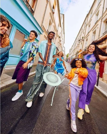 A group of diverse young adults walking down a narrow city street, wearing colorful casual clothing, holding a small chair, and smiling.