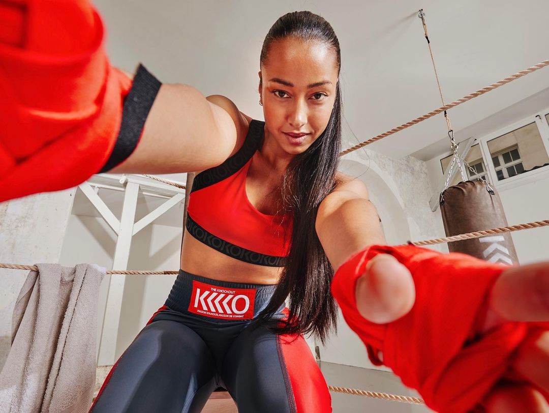 Female boxer in sports bra and leggings poses with wrapped fists inside a boxing ring, looking directly at the camera.