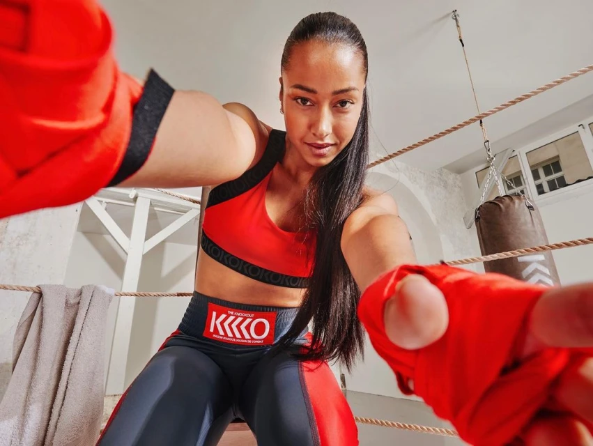 Female boxer in sports bra and leggings poses with wrapped fists inside a boxing ring, looking directly at the camera.