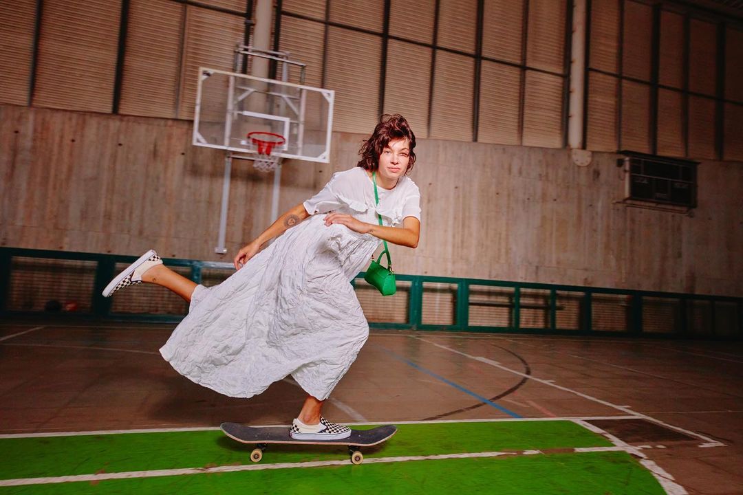 Woman in white dress and Vans rides skateboard indoors in a gymnasium by basketball hoop, in a dynamic fashion and sports lifestyle image.