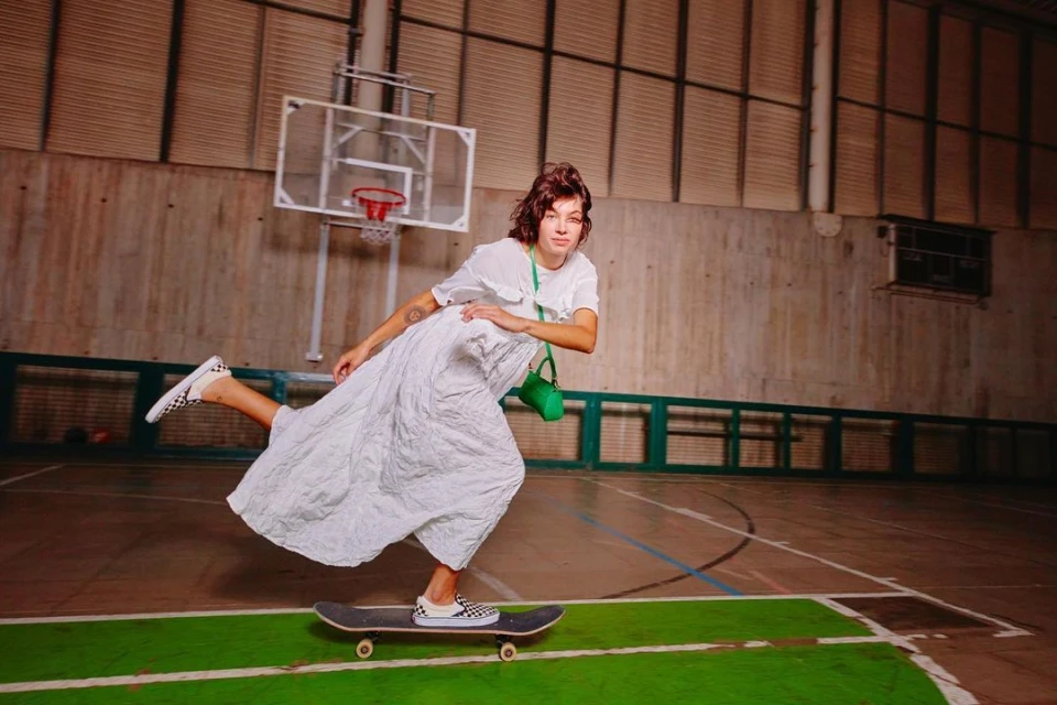 Woman in white dress and Vans rides skateboard indoors in a gymnasium by basketball hoop, in a dynamic fashion and sports lifestyle image.