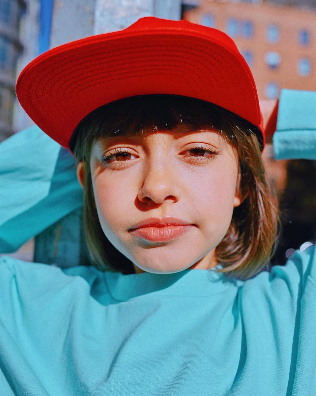 Close-up of a young woman with neutral expression, wearing a red baseball cap and turquoise long-sleeved shirt, posing outdoors against a building.
