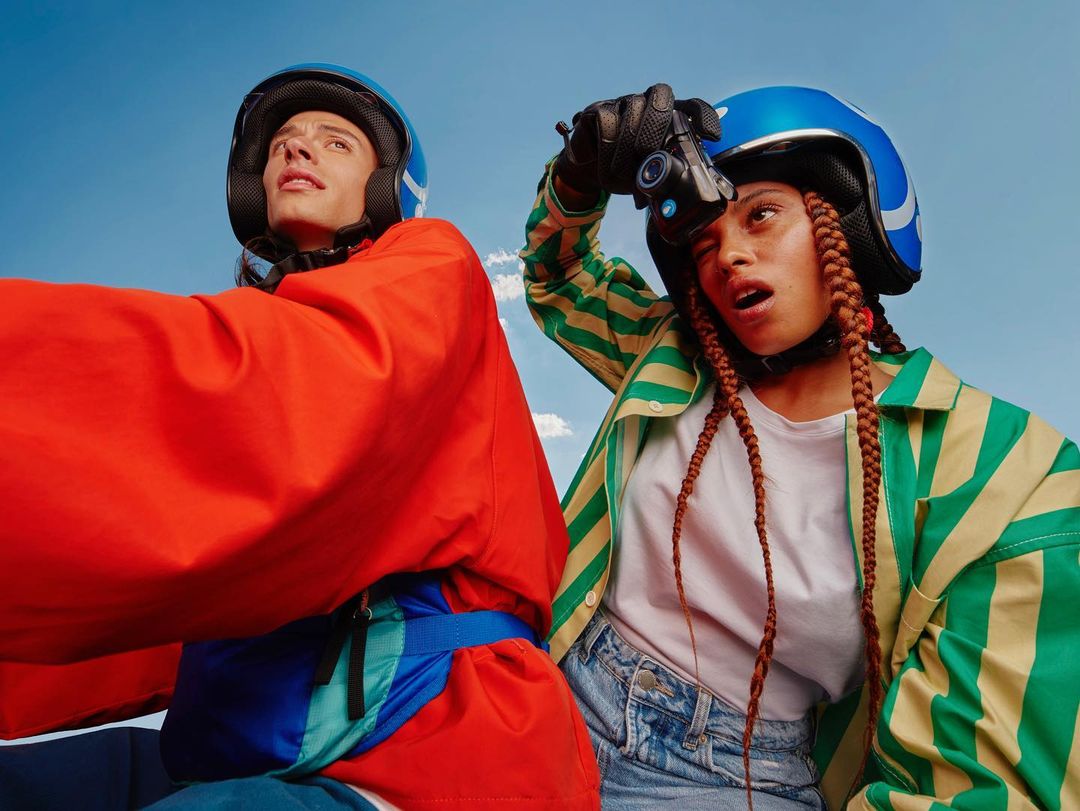 Two people wearing helmets and colorful jackets look upward against a bright blue sky with soft clouds, captured in a wide outdoor shot.