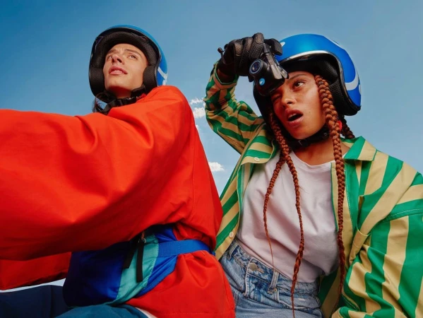 Two people wearing helmets and colorful jackets look upward against a bright blue sky with soft clouds, captured in a wide outdoor shot.