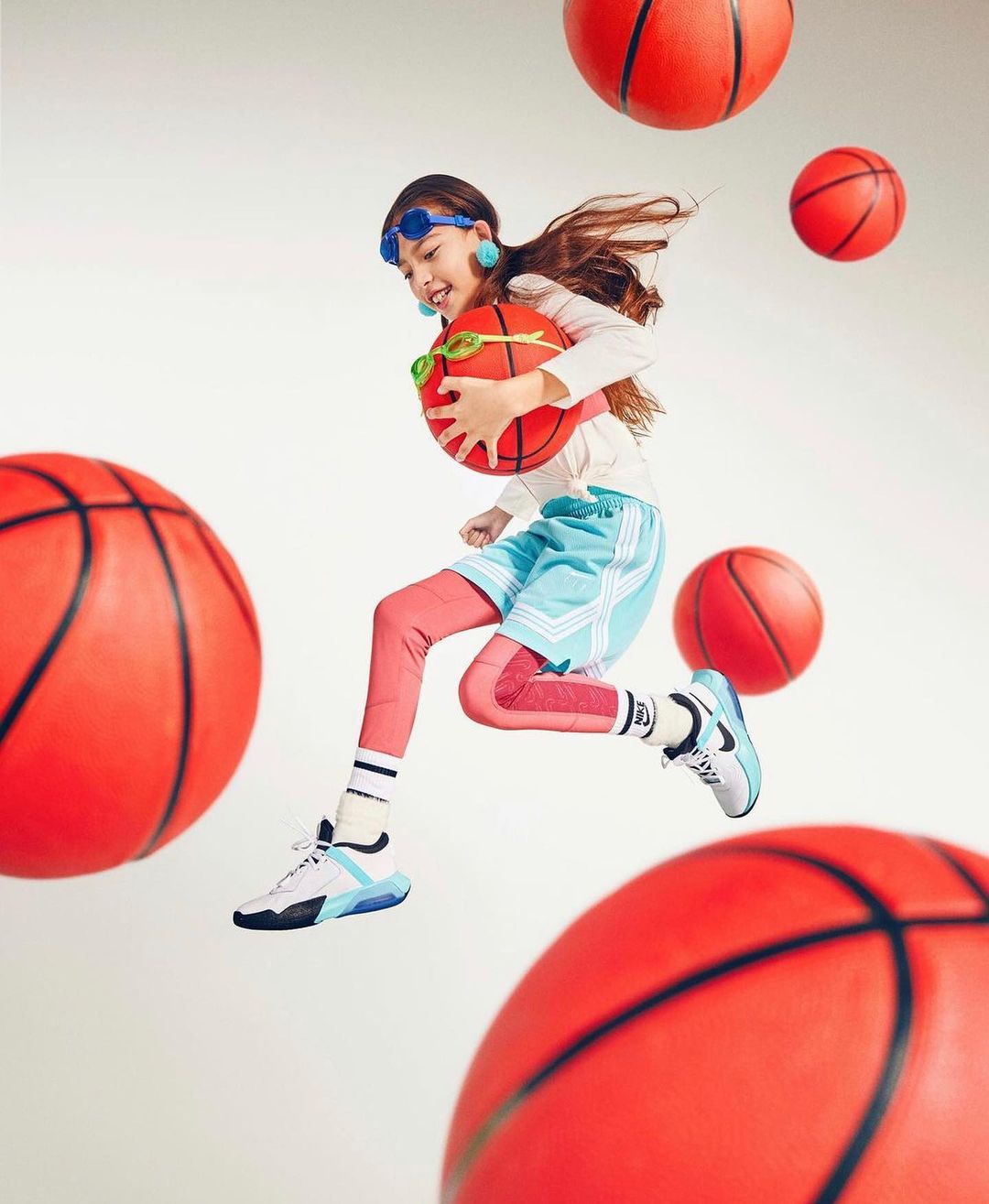 A girl in sports attire mid-jump with a basketball, surrounded by floating basketballs against a bright, neutral background.